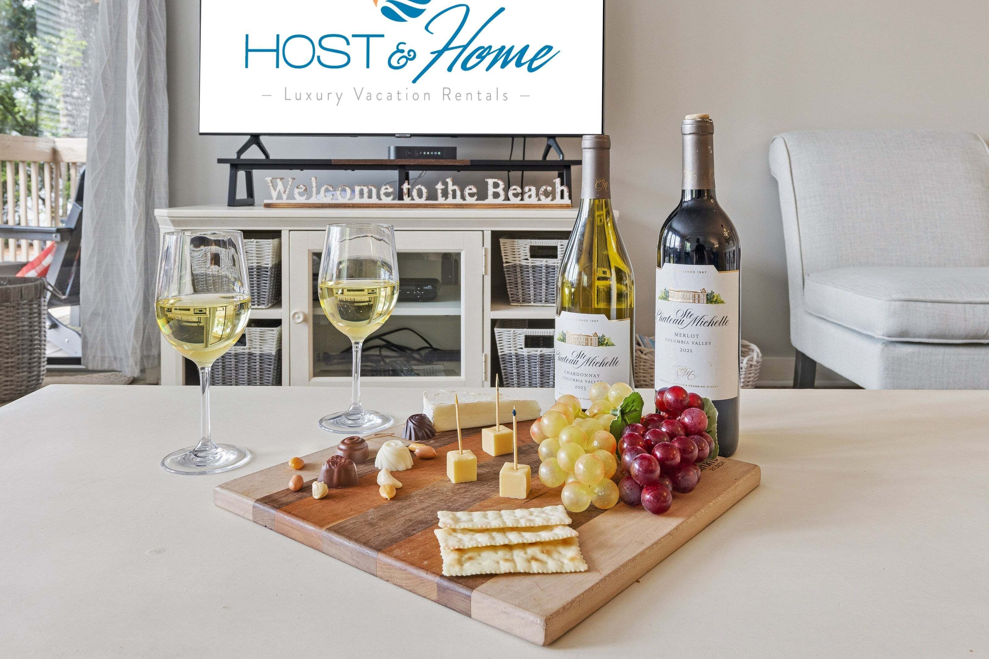 Taula amb una ampolla de vi, gots, formatge i raïms, amb un cartell que diu 'HOST & Home - Luxury Vacation Rentals' al fons.