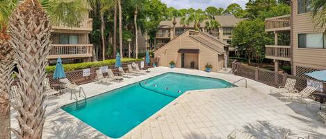 A swimming pool area with lounge chairs and umbrellas, surrounded by palm trees and residential buildings.