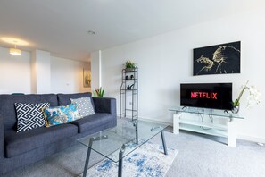 Een moderne woonkamer met een grijze bank, een glazen salontafel, een televisie op een dressoir en een zwart kunstwerk aan de muur.