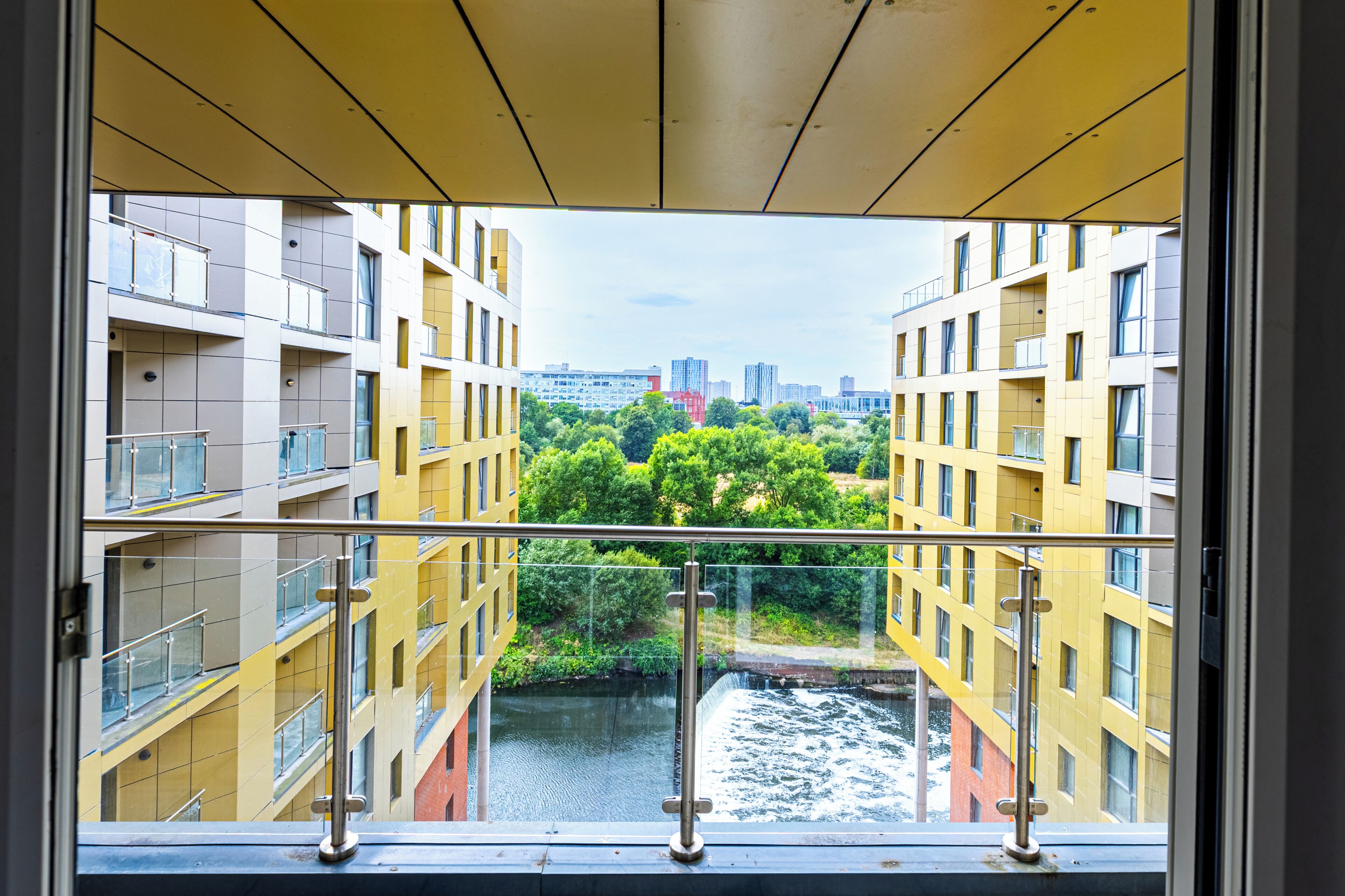 A view through a window of a yellow building with balconies, overlooking a river and a cityscape.