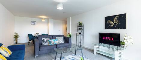 A modern living room with a grey sofa, a blue sofa, a glass coffee table, a TV on a stand, and a wall-mounted shelf with a plant.