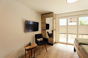 A modern room with a flat-screen TV mounted on the wall, a wooden side table with a remote control, a black chair with a cushion, and a bed with a green blanket. - The Art of Alpine Living, Wallis 1097 (Wallis)