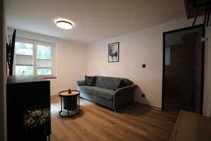 A living room with a grey sofa, a round wooden table, and a window with blinds. - Mornings & Refined Alpine Living 1098 (Wallis)