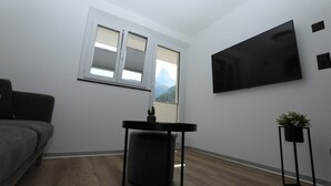 Ein Zimmer mit grauem Sofa, einem kleinen Tisch mit Pflanze, einem an der Wand montierten Fernseher und einem Fenster mit Bergblick.