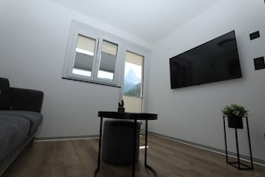 A room with a grey sofa, a small table with a plant, a TV mounted on the wall, and a window with a view of mountains. - Alpine Calm with Unforgettable Views 1099 (Wallis)