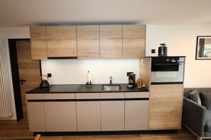 A compact kitchen with a black countertop, wooden cabinets, a built-in oven, and a coffee machine. - Alpine Calm with Unforgettable Views 1099 (Wallis)
