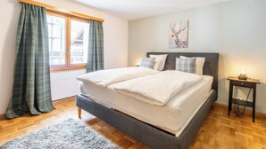 Un dormitorio con cama, mesita de noche, ventana con cortinas y un cuadro en la pared.