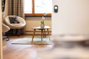 Ein Zimmer mit Holzboden, einem Sessel, einem kleinen Tisch mit Pflanze und einem Fenster mit Vorhängen.