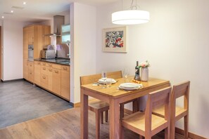 Ein Essbereich mit Holztisch und Stühlen, im Hintergrund eine moderne Küche und eine Pendelleuchte über dem Tisch.