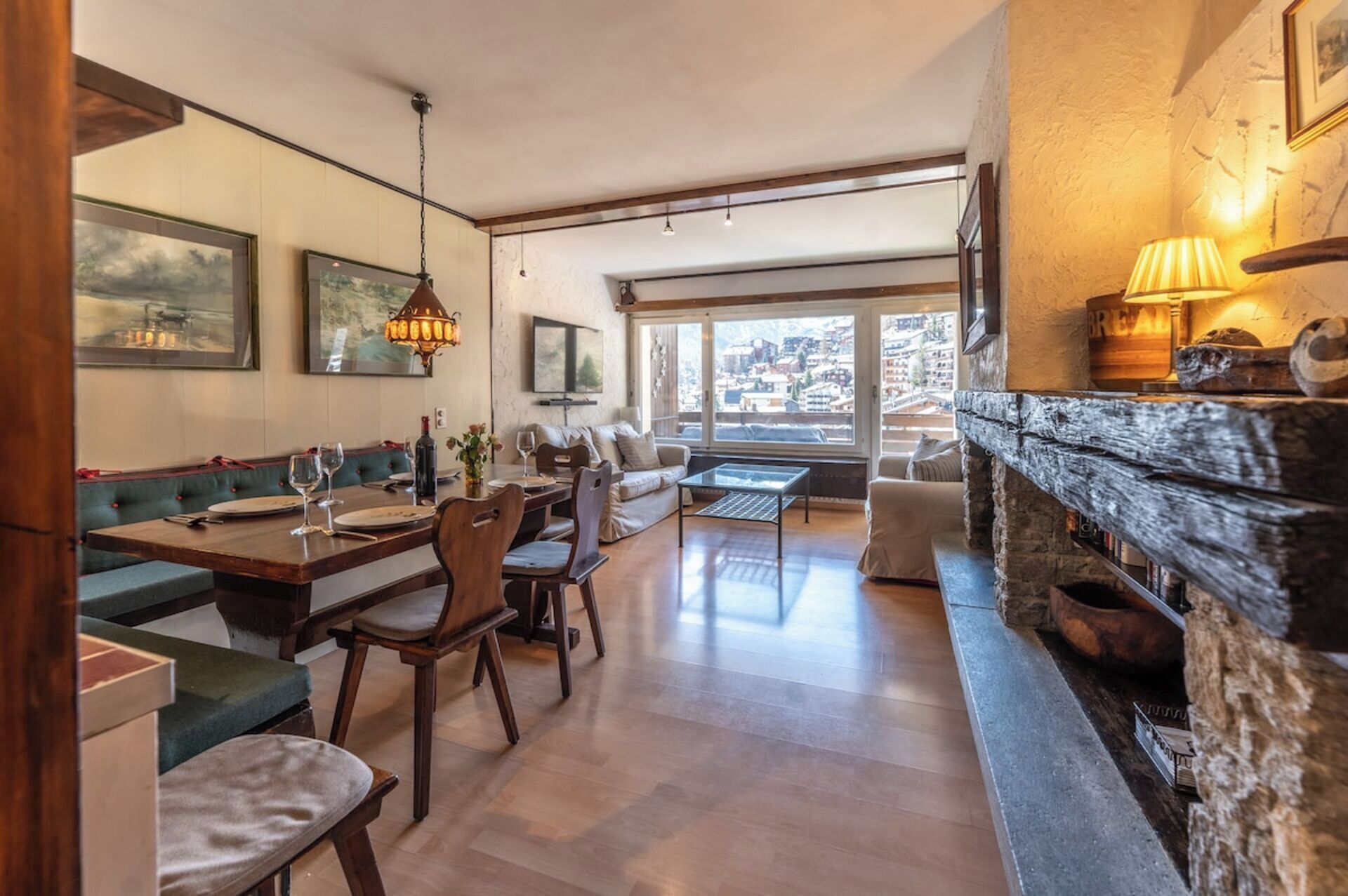 A dining area with a wooden table, chairs, and a bench. There is a fireplace with a stone wall and a lamp.