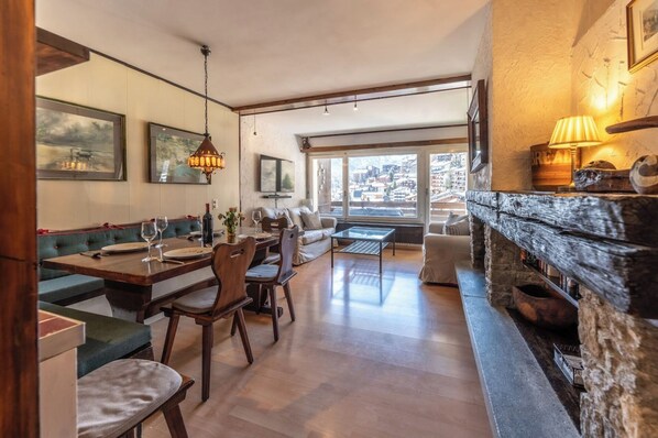 Un coin repas avec une table en bois, des chaises et un banc. On y trouve une cheminée avec un mur en pierre et une lampe.