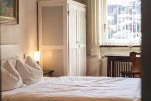 Ein Hotelzimmer mit Bett, Schrank, Nachttisch mit Lampe und Fenster mit Blick auf Gebäude.