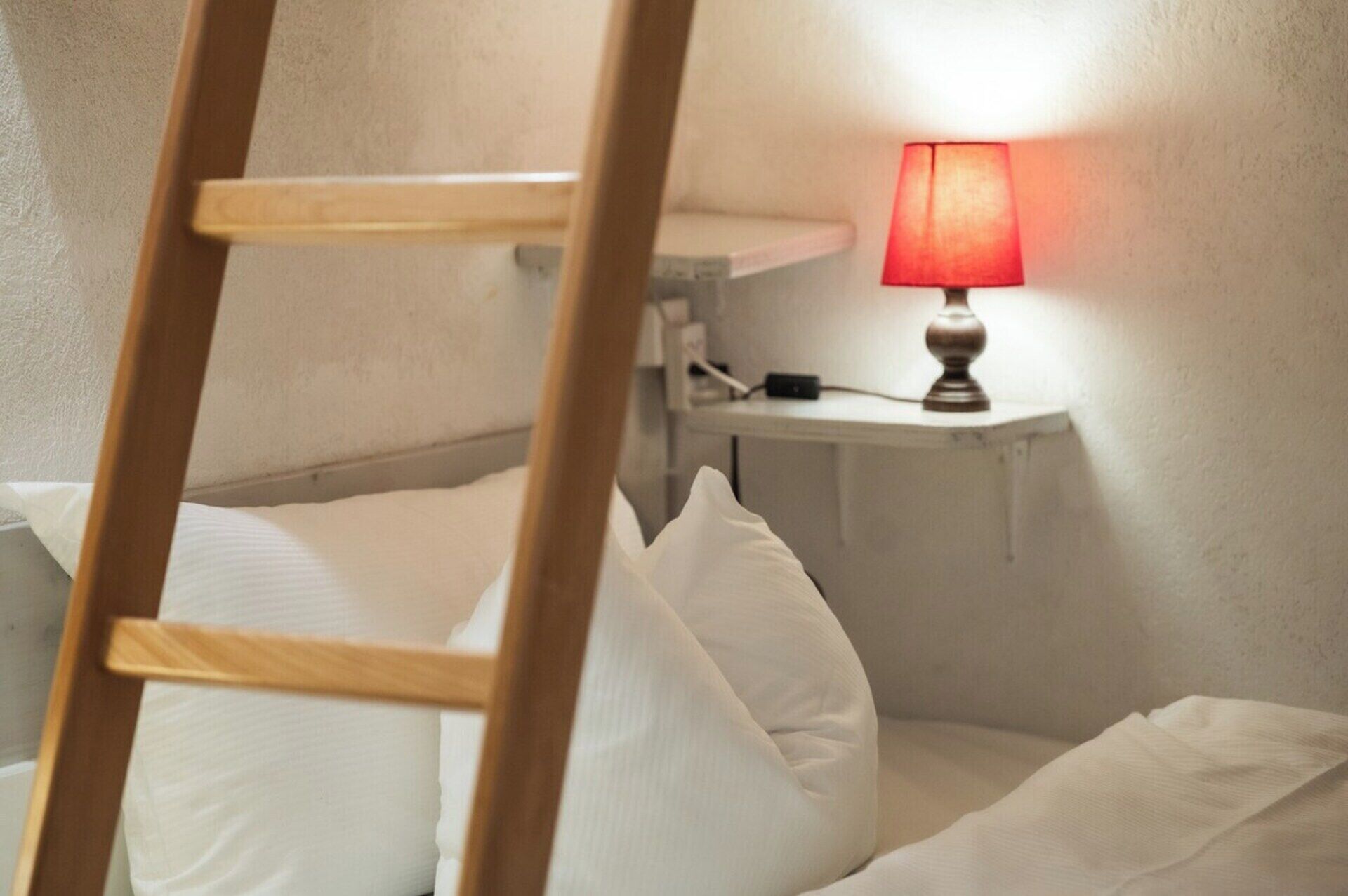 Ein Stockbett mit Holzlade, ein Nachttisch mit roter Lampe und weißer Bettwäsche.