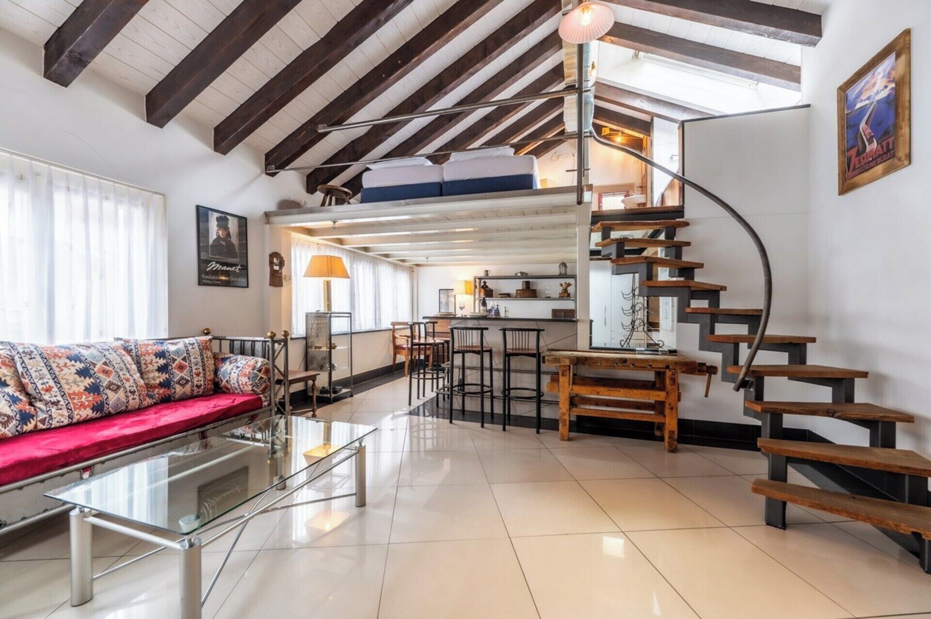 A modern living space with a glass coffee table, a sofa with patterned cushions, a kitchen area with a bar, and a spiral staircase.