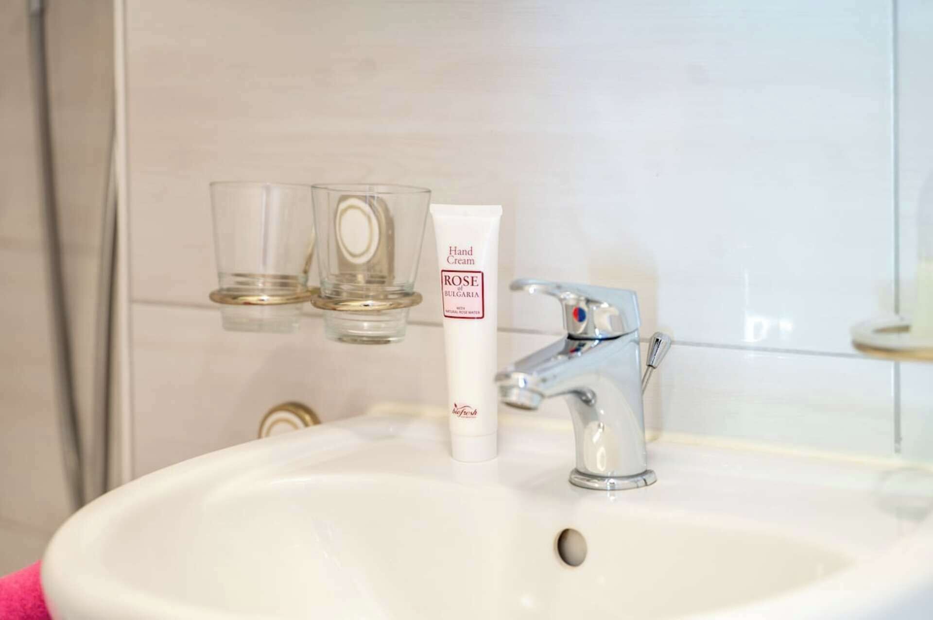 A bathroom sink with a hand cream tube labeled 'ROSE of Bulgaria' and two clear glasses on a shelf.