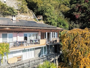 A building with a balcony overlooking a pool, surrounded by trees and a stone wall. - Your Elegant  Mountain Retreat 1103 (Ticino)