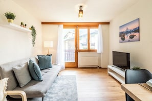Ein Wohnzimmer mit Sofa, Fernseher, Esstisch und einem Fenster mit Vorhängen.