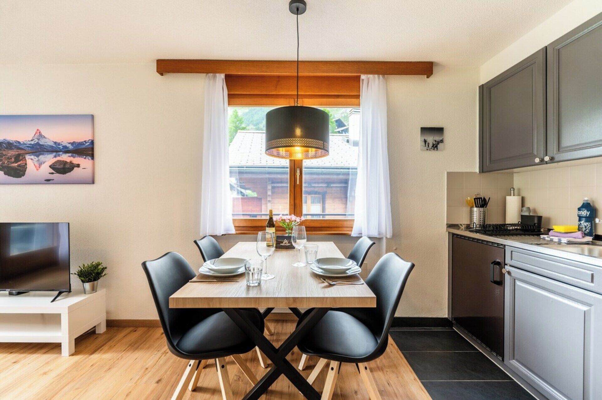 Un coin repas avec une table en bois, des chaises noires, un luminaire suspendu et une vue sur une montagne par la fenêtre.