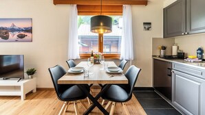 A dining area with a wooden table, black chairs, a hanging light fixture, and a view of a mountain through a window. - Sophisticated Alpine Living with Views 1091 (Wallis)