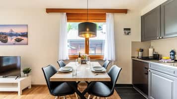 Un coin repas avec une table en bois, des chaises noires, un luminaire suspendu et une vue sur une montagne par la fenêtre.