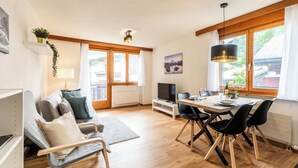 A modern living room with a sofa, a dining table with chairs, a TV, and a large window with curtains. - Sophisticated Alpine Living with Views 1091 (Wallis)