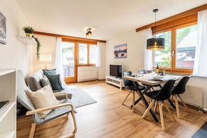 Ein modernes Wohnzimmer mit einer Couch, einem Esstisch mit Stühlen, einem Fernseher und einem großen Fenster mit Vorhängen.