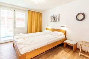 A double bed with wooden headboard and footboard, a bedside table, a bench, a window with curtains, and a wall-mounted mirror. - Effortless Alpine Living 1411 (Valais)