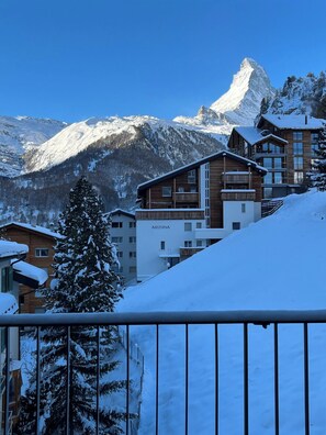 A snow-covered mountain resort with a prominent peak in the background, wooden buildings with balconies, and a snow-laden pine tree in the foreground. - Effortless Alpine Living 1411 (Valais)