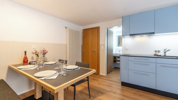 A modern kitchen with blue cabinets, a wooden dining table set for four, and a view into the adjacent dining area. - A Beautiful Alpine Escape in Zermatt 1092 (Wallis)