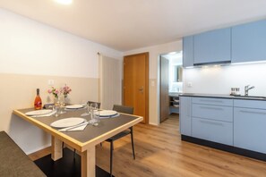 A modern kitchen with blue cabinets, a wooden dining table set for four, and a view into the adjacent dining area. - A Beautiful Alpine Escape in Zermatt 1092 (Wallis)
