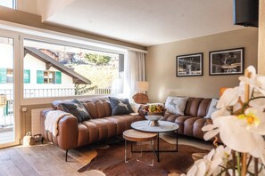 Un salon avec un canapé en cuir marron, une table basse ronde et une vue sur un immeuble par le biais d’une grande fenêtre.