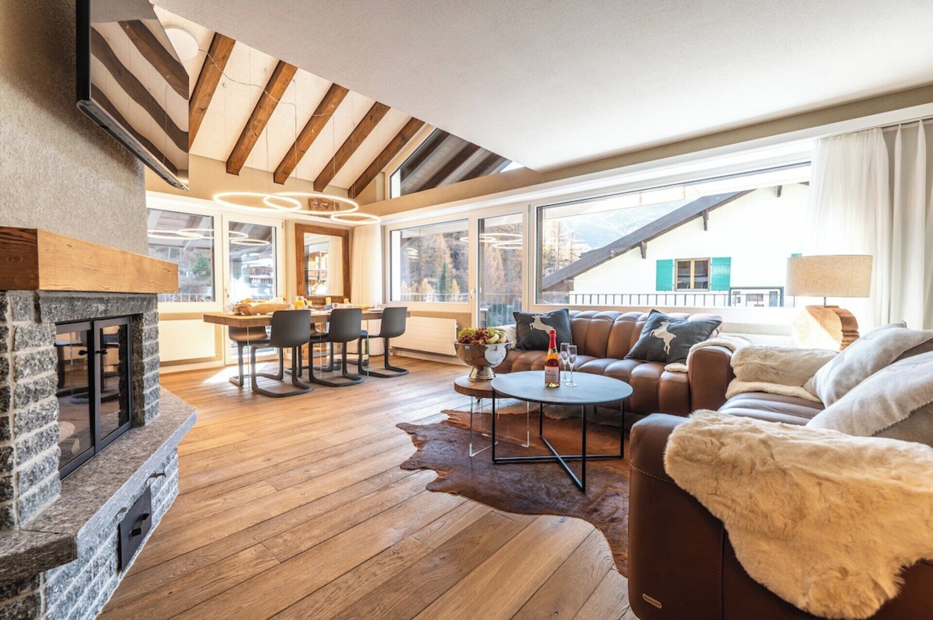 Ein modernes Wohnzimmer mit Kamin, Holzboden, Ledersofa und großen Fenstern mit Blick auf die Berge.
