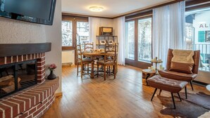 Un salon chaleureux avec une cheminée, une table à manger en bois, des sièges confortables et de grandes fenêtres donnant sur l’extérieur.