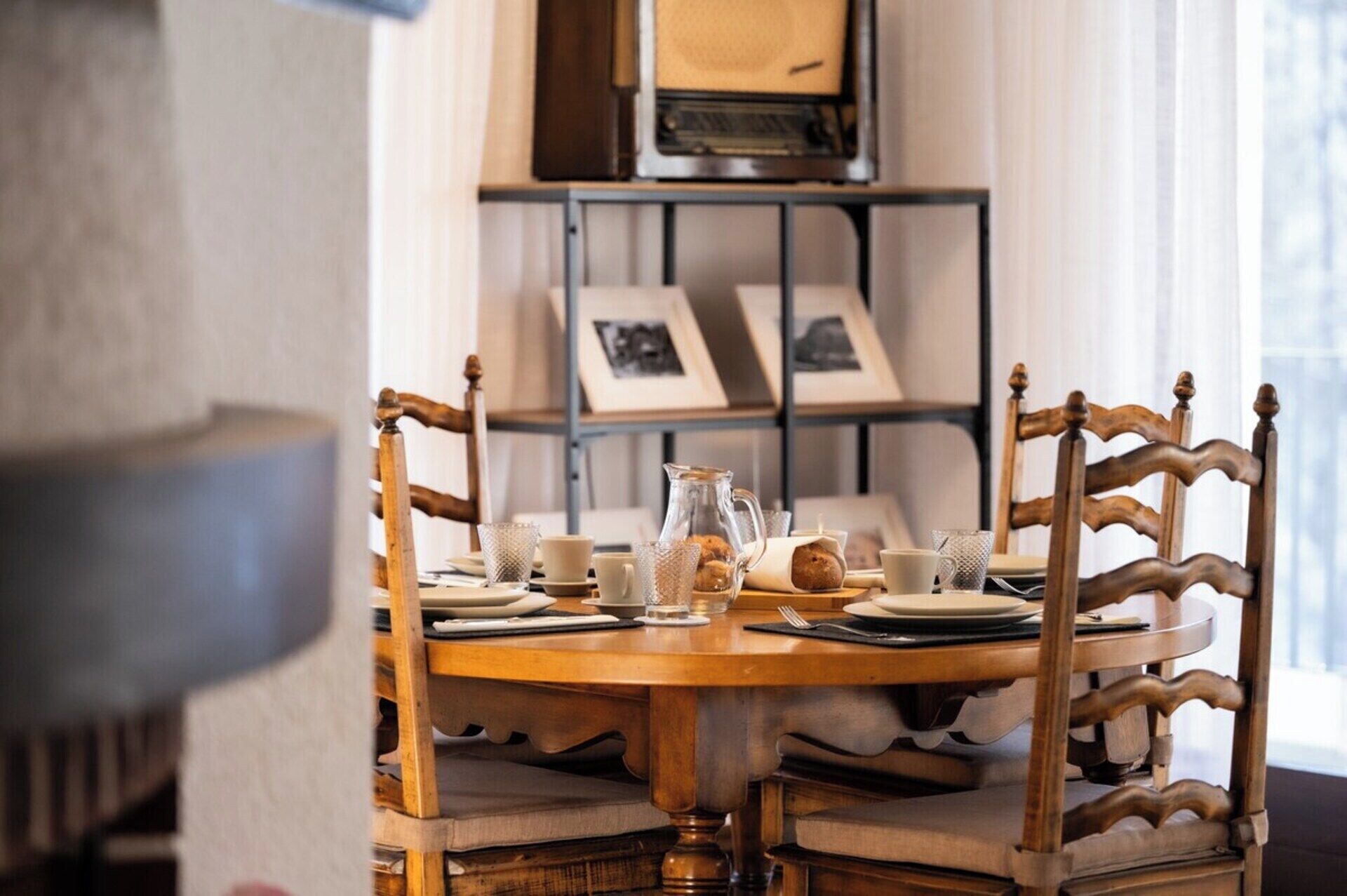 A dining table set with chairs, a pitcher, and plates, with a bookshelf in the background.