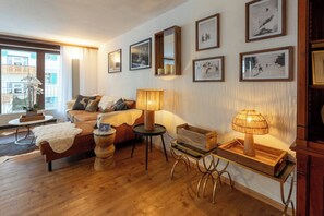 Ein gemütliches Wohnzimmer mit einer Couch, einem Couchtisch und einer Lampe. An den Wänden hängen Bilderrahmen und ein Fenster mit Vorhängen.