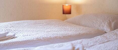 A neatly made bed with a white blanket and pillows, a wall-mounted lamp, and folded towels on the bed.