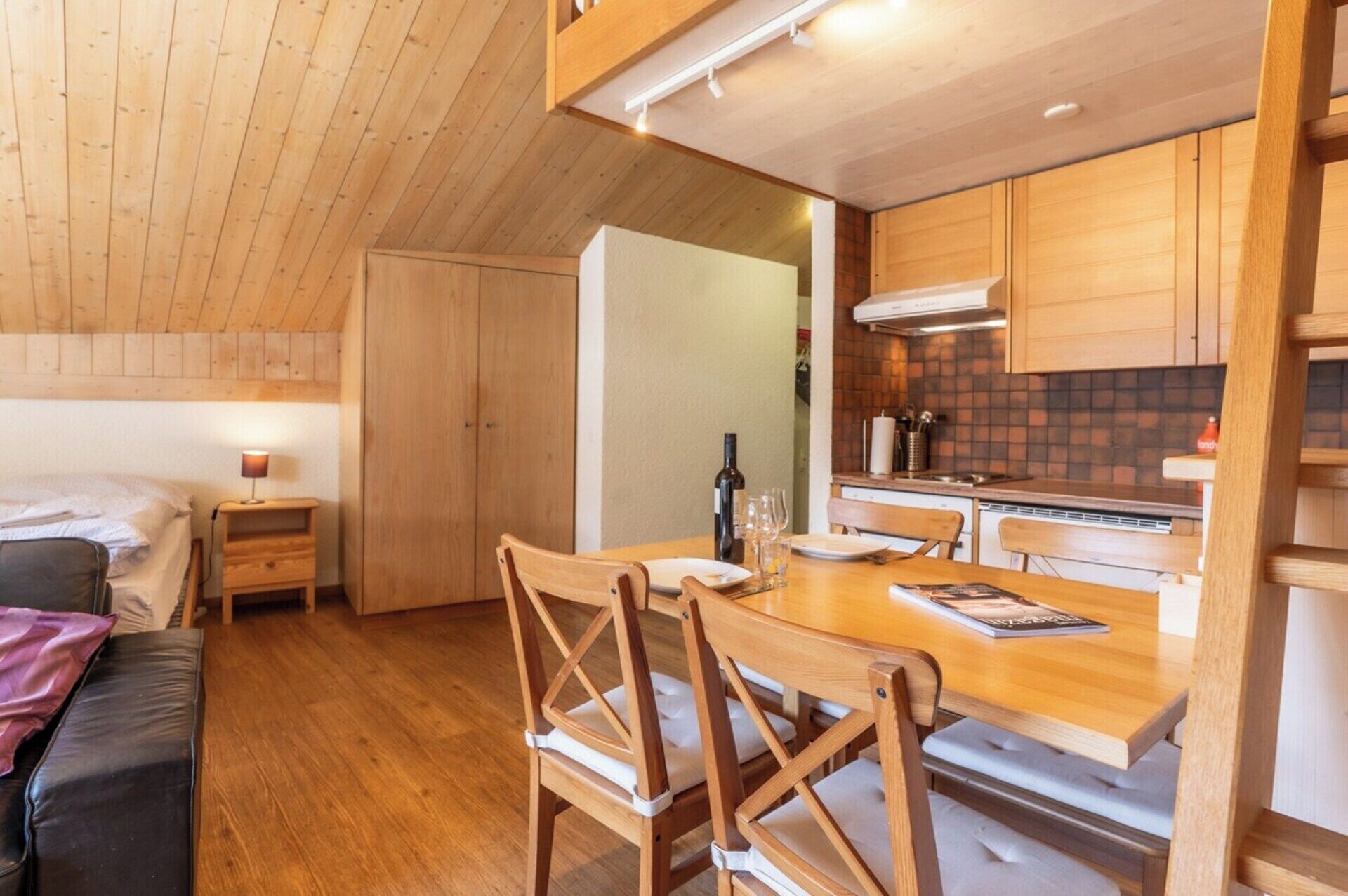 A compact room with a bed, a wooden dining table with chairs, a kitchen area with wooden cabinets, and a small bedside table with a lamp.