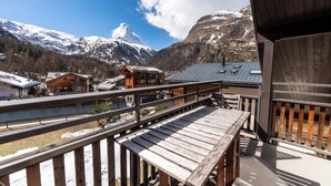 A wooden deck with a view of snow-capped mountains and a chalet-style building. - Serene Alpine Luxury in Zermatt 1089 (Wallis)