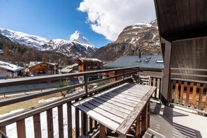 Eine Holzterrasse mit Blick auf schneebedeckte Berge und ein Chalet-ähnliches Gebäude.