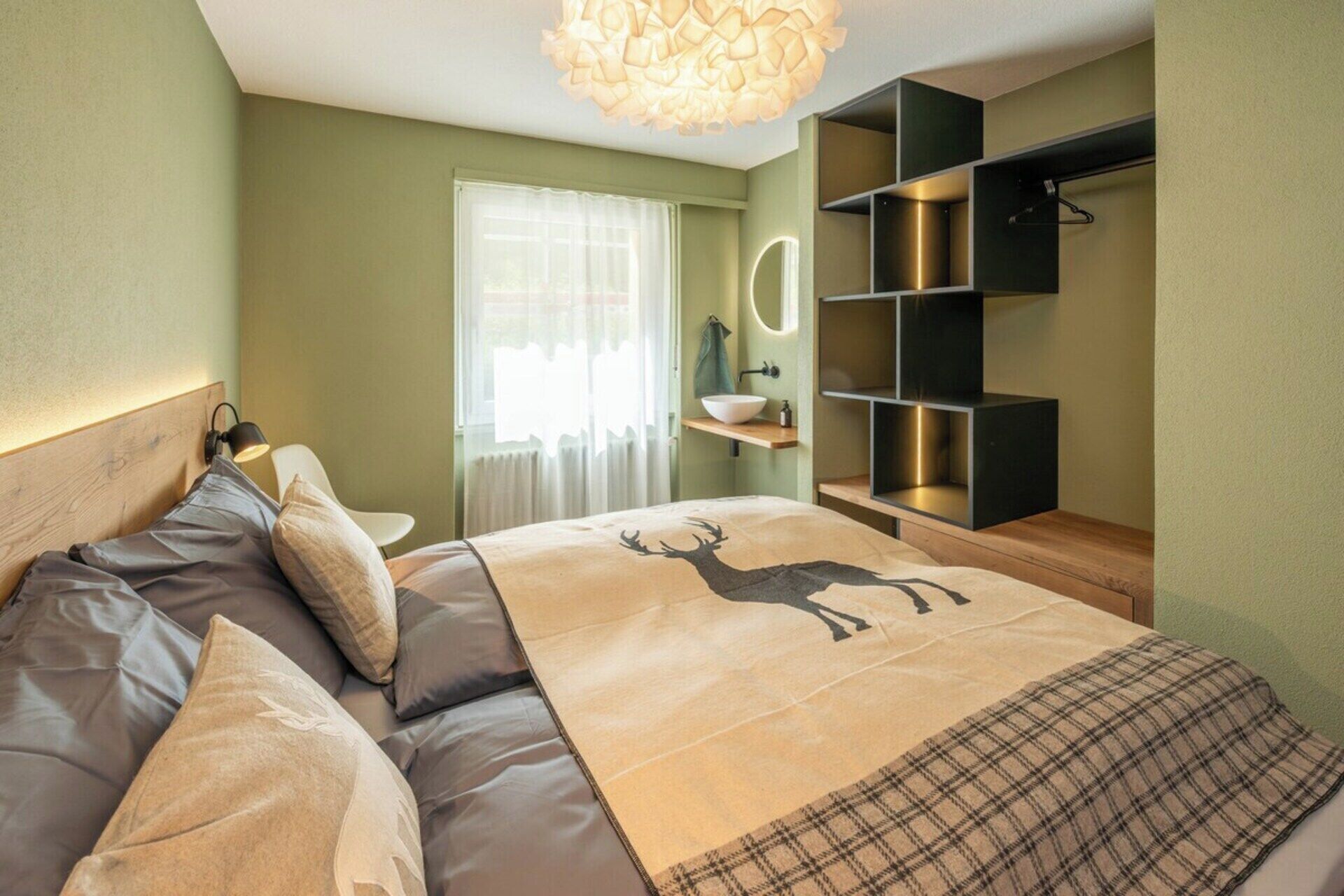 A bedroom with a bed, a bedside table, a mirror, a shelf unit, and a hanging light fixture.