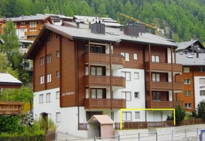 Un hôtel à plusieurs étages doté de balcons, avec un télésiège visible au loin et un paysage montagneux en arrière-plan.