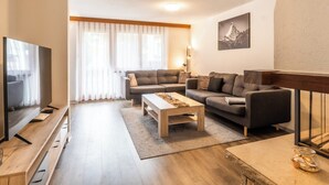 Uma sala de estar moderna com televisão de tela plana, dois sofás, uma mesa de centro e uma janela grande com cortinas leves.