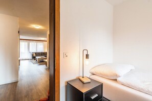 Ein Hotelzimmer mit einem Bett, einem Nachttisch mit Lampe, Holzboden und Blick auf den Wohnbereich durch eine geöffnete Tür.