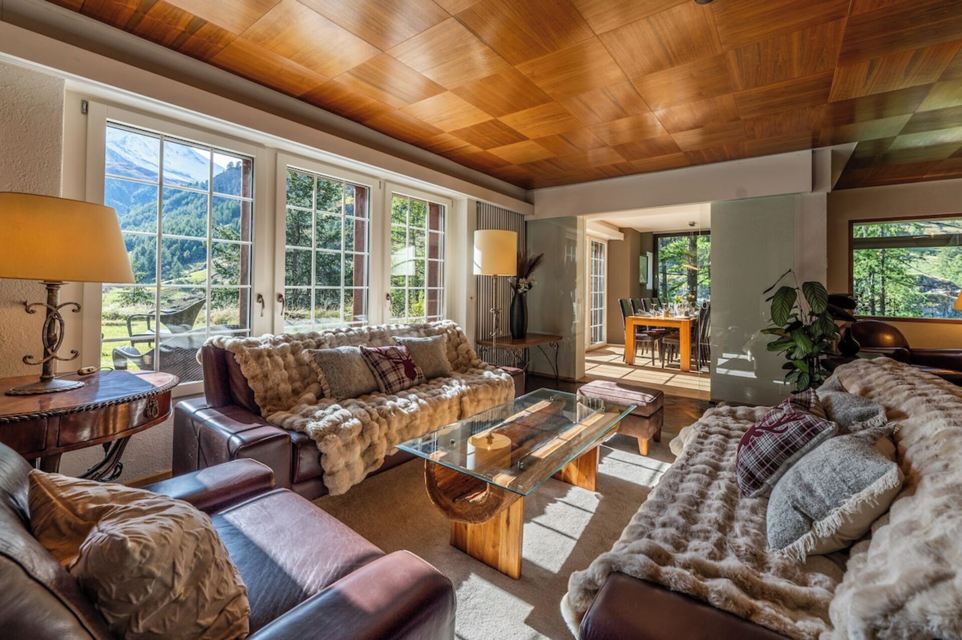 A spacious living room with a wooden ceiling, leather sofas, a glass coffee table, and large windows offering a view of the outdoors.