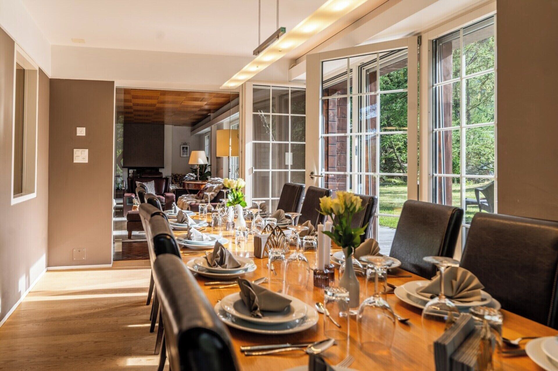 A dining area with a long table set for a meal, large windows, and a view of the outdoors.