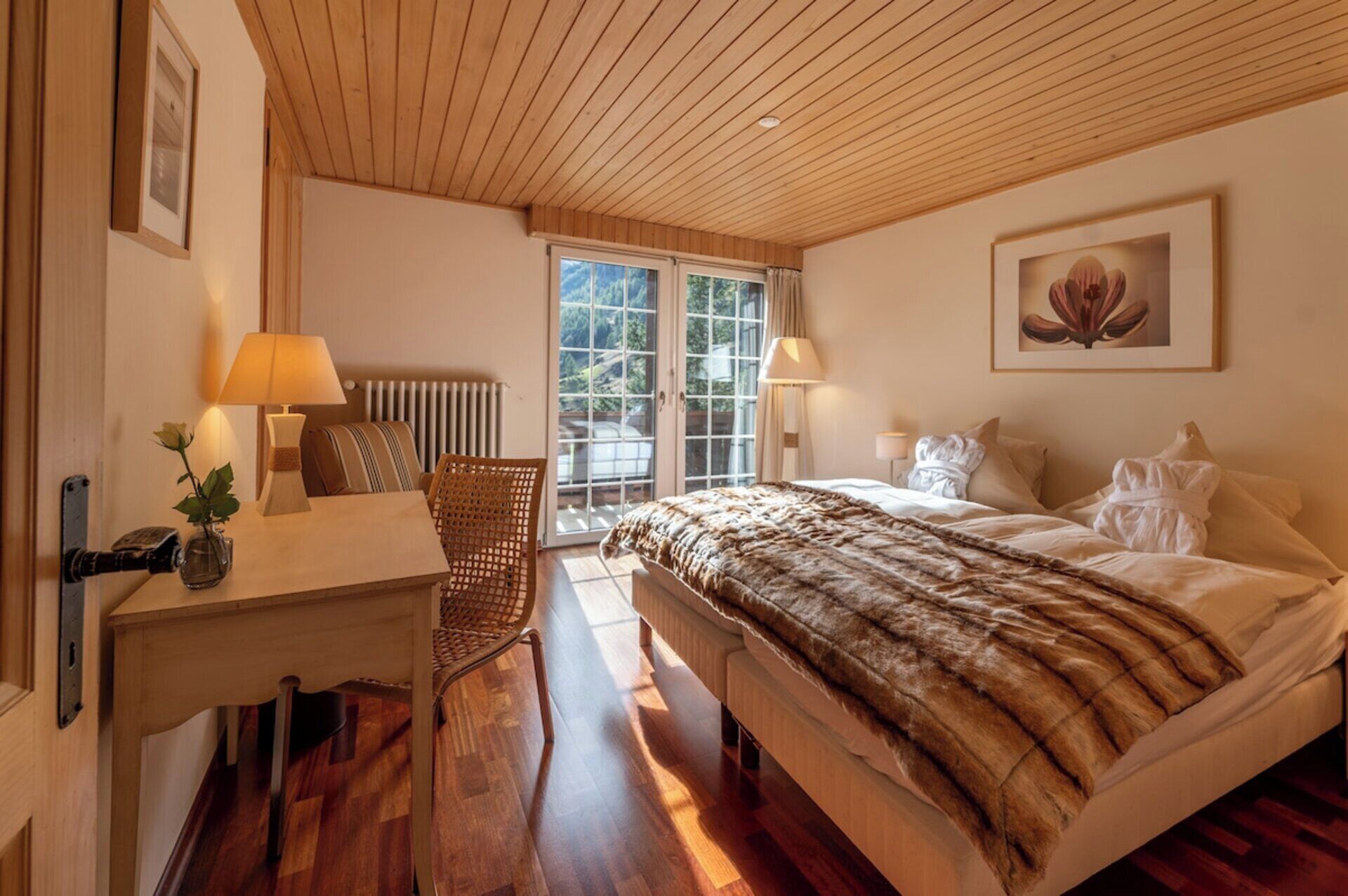 A bedroom with a wooden ceiling, a bed with a fur-like cover, a desk with a lamp, and a chair.