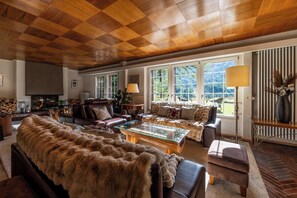 A cozy living room with a wooden ceiling, leather sofas, a glass coffee table, and large windows offering a view of the outdoors.