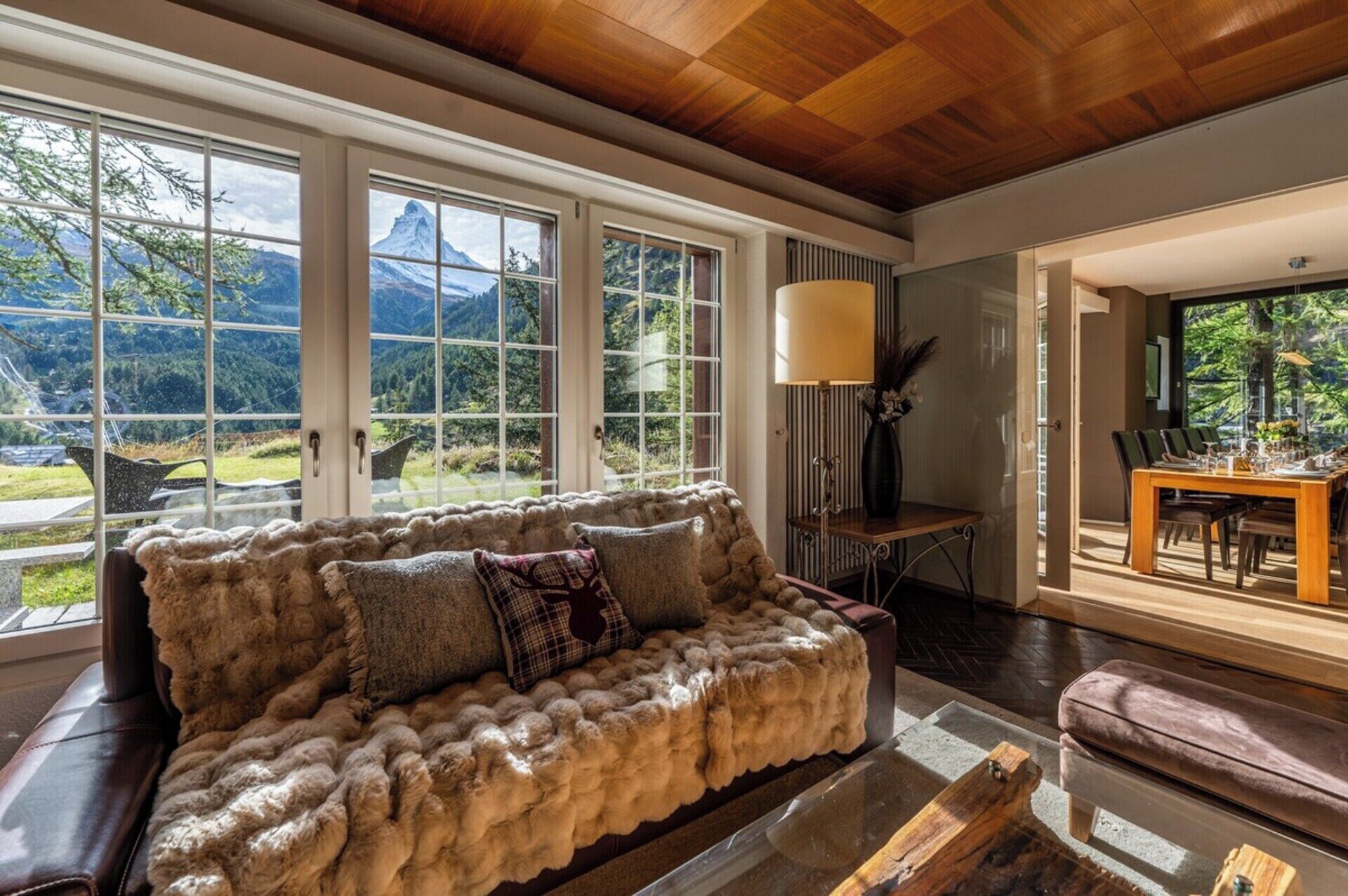Ein gemütliches Wohnzimmer mit einem fellbezogenen Sofa, großen Fenstern mit Blick auf die Berge, einem Essbereich mit Tisch und Stühlen sowie einer Lampe.
