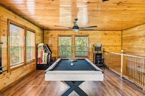 A pool table in a wooden room with a ceiling fan, arcade games, and large windows. - NEW! Modern Smokies Cabin! Hot Tub! Games! POOL! (Pigeon Forge)