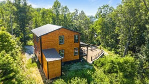 A wooden cabin with a deck, surrounded by dense forest. - NEW! Modern Smokies Cabin! Hot Tub! Games! POOL! (Pigeon Forge)